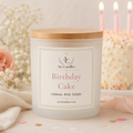 A frosted white Arc Candles 'Birthday Cake' scented candle with a light wood lid, set on a table with a blurred birthday cake, lit candles, and flowers in the background.
