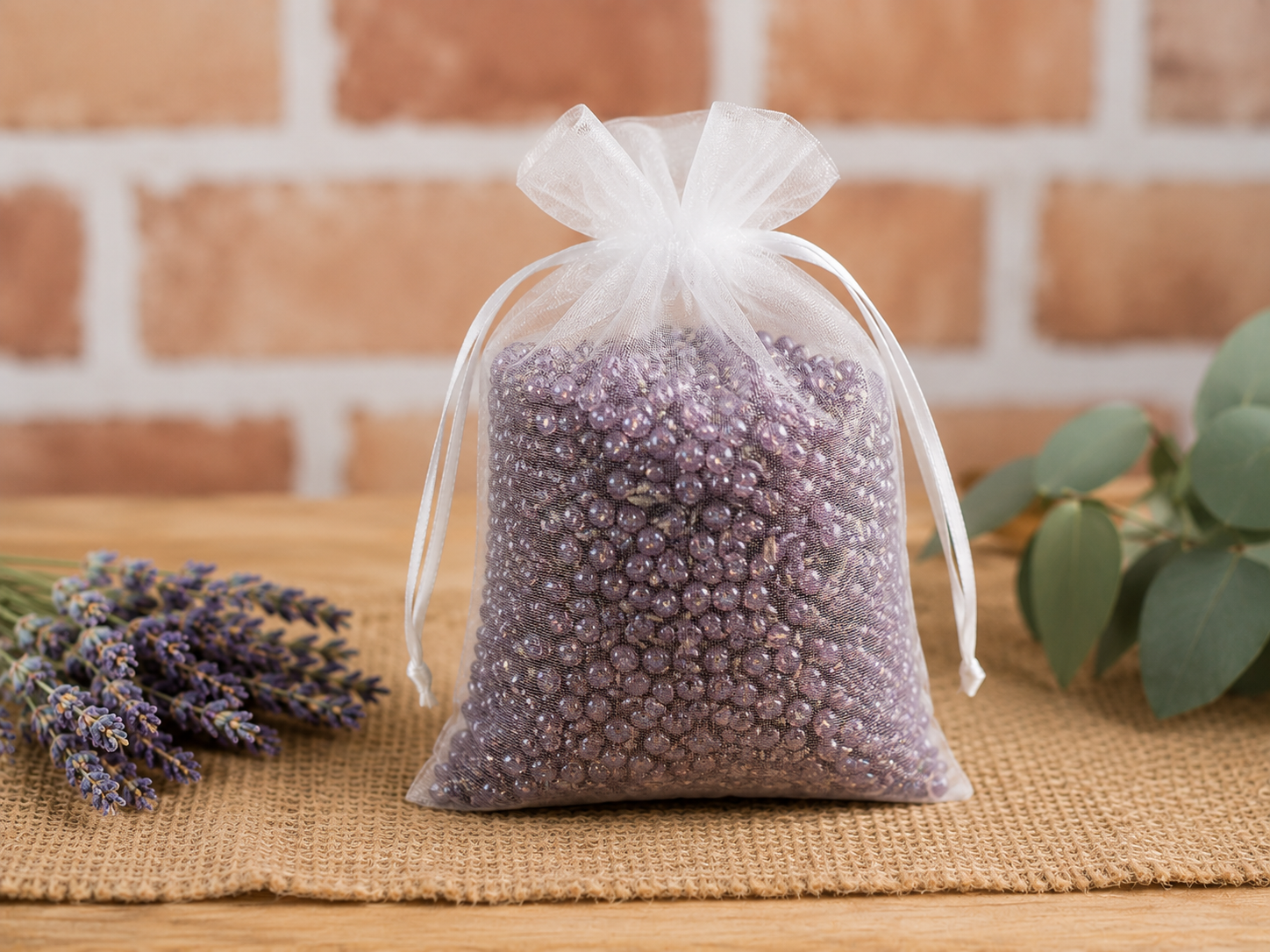 A clear organza drawstring sachet bag filled with lavender beads, styled on a burlap mat with lavender sprigs and green leaves against a brick wall background.
