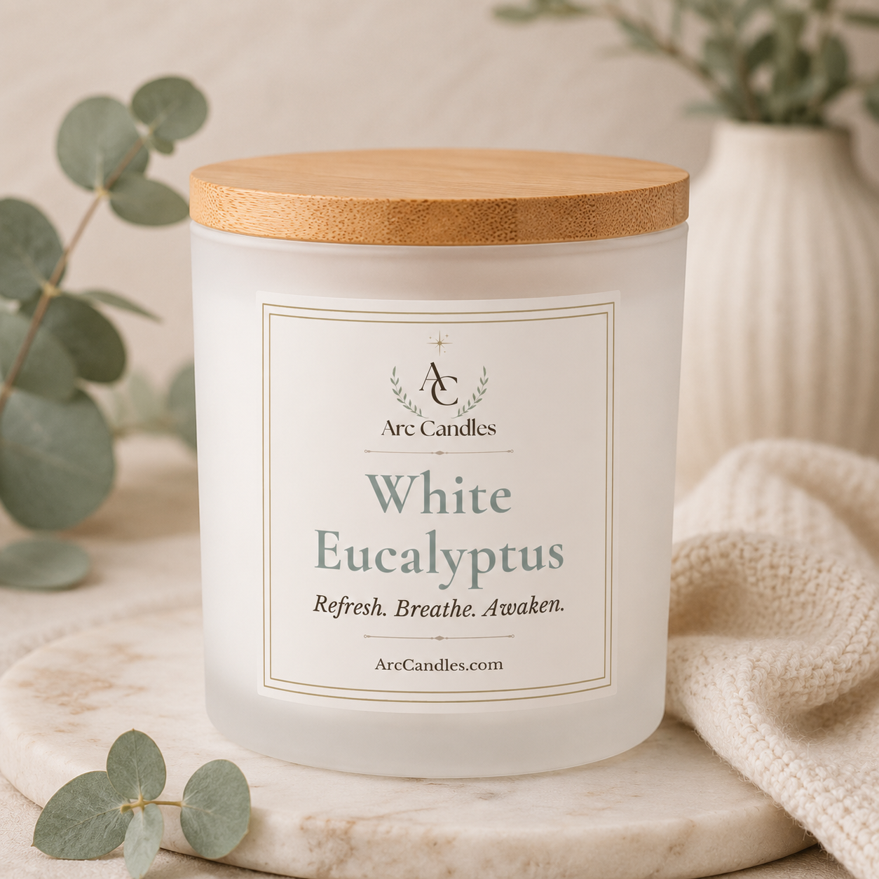 A frosted glass jar candle with a light wood lid sits on a marble surface in a softly styled setting. The front label reads Arc Candles and White Eucalyptus, with eucalyptus leaves and a cream knit fabric in the background.