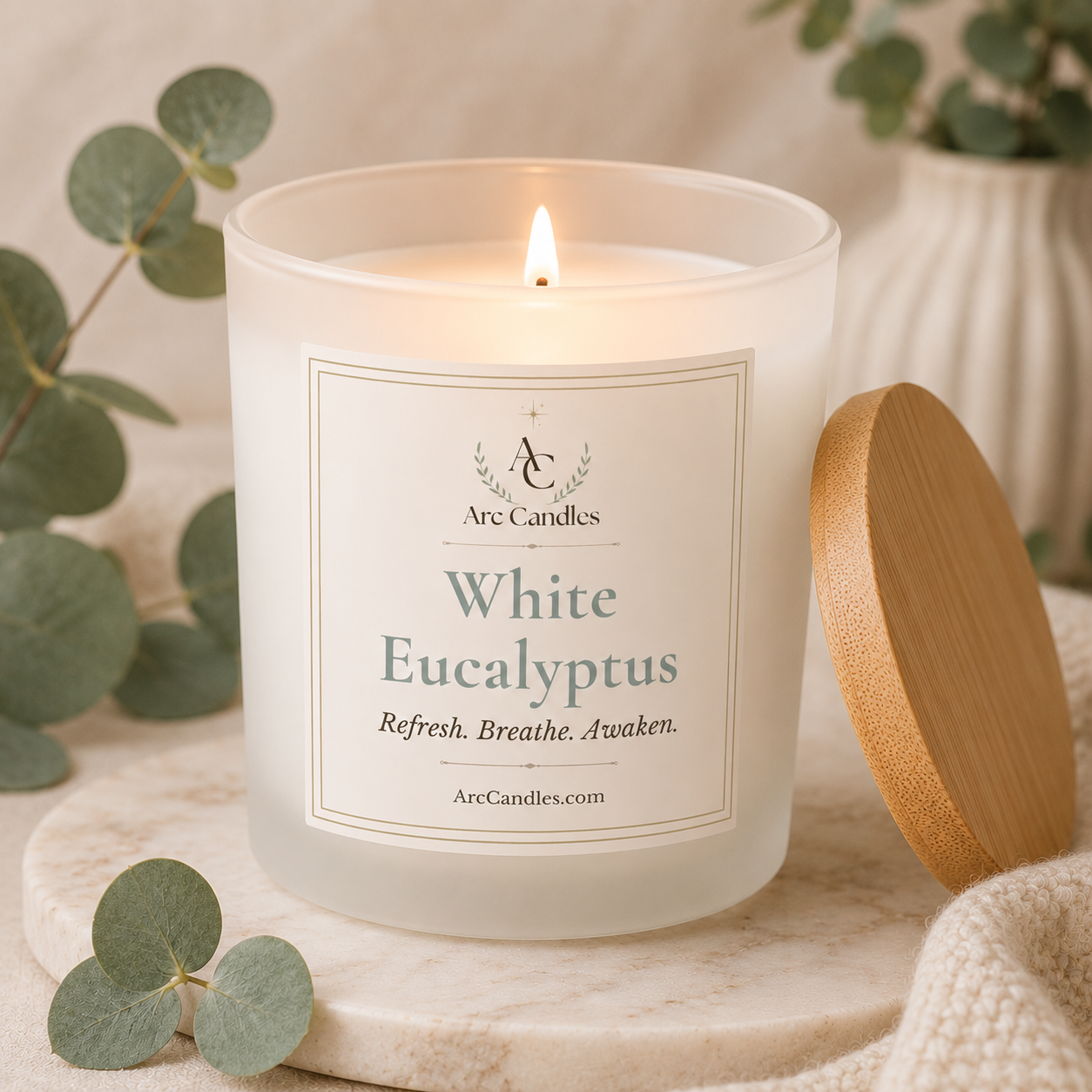 A lit white soy candle in a frosted glass jar with a wooden lid resting beside it on a marble surface. The front label reads Arc Candles and White Eucalyptus, with eucalyptus leaves and a soft neutral background.